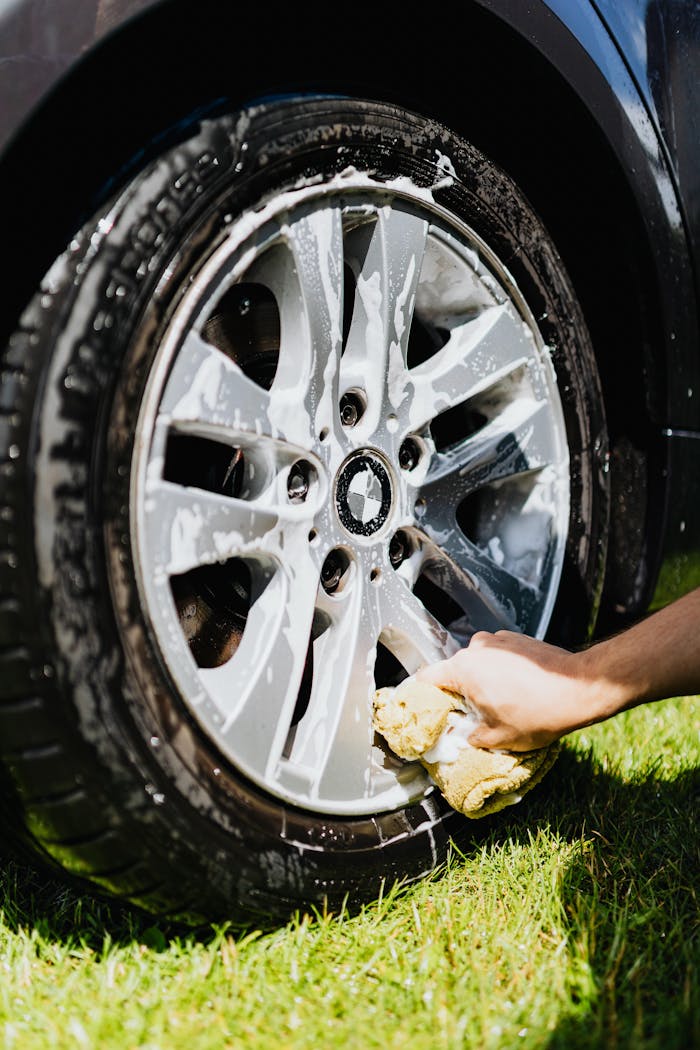 about-us Close-up of a car wheel with soap suds and hand cleaning it on grass.