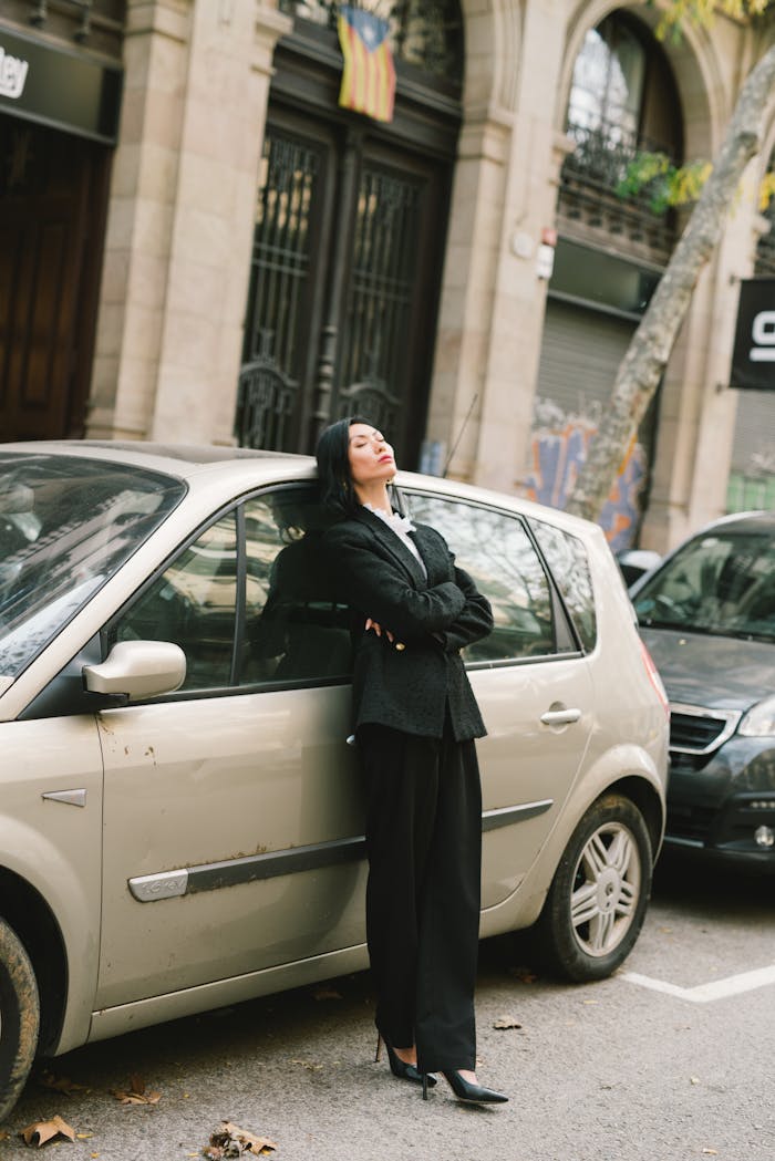 Woman in black coat leaning on car on city street, arms crossed, looking relaxed.